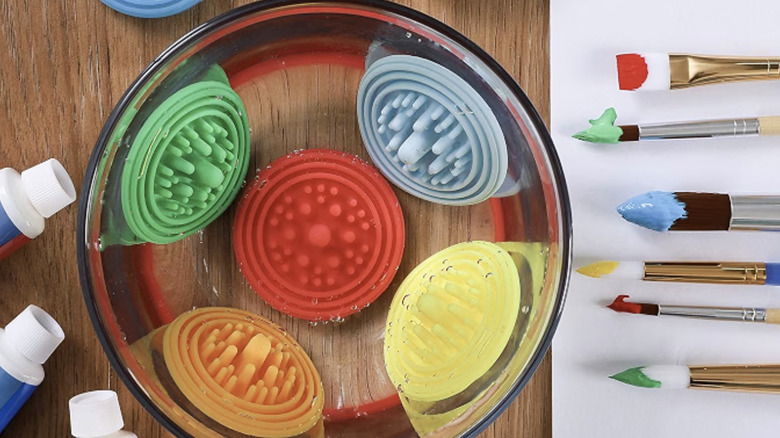 Five different colored paint pucks in a bowl of water next to brushes with paint