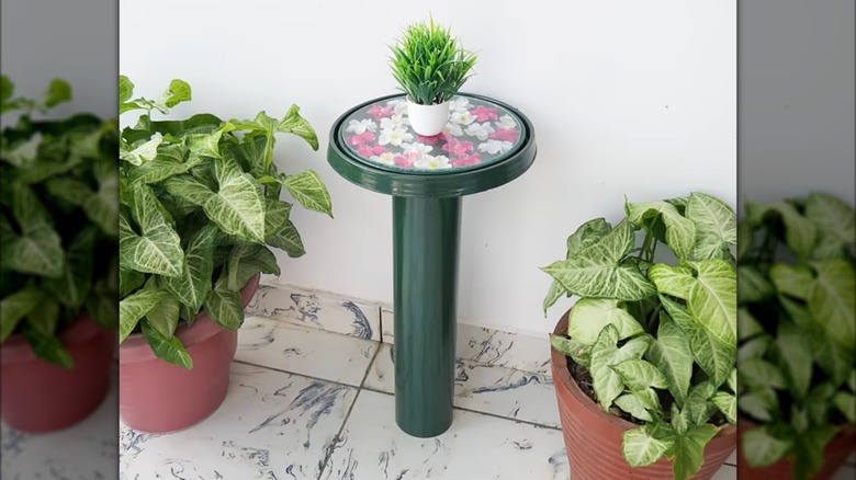A DIY side table made using a bucket lid is flanked by two potted plants in a living room.