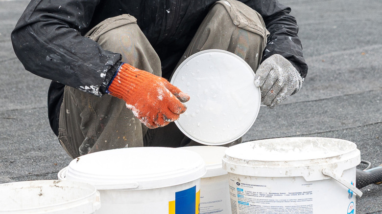 A man holds the lid of a white paint bucket in his gloved hands with more buckets in front of him.