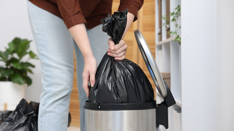 A person removing a full trash bag from a metal trash can.