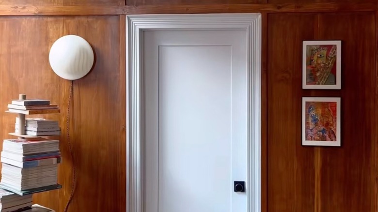 A white door surrounded by DIY mid-century modern wood paneling styled with a bookshelf, wall light, and picture frames