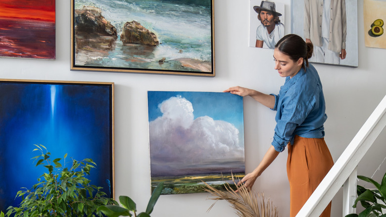A woman hangs artwork on a wall full of framed canvases