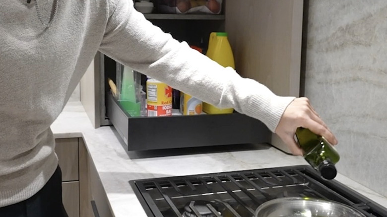 A man pours oil into a pan on a stovetop with a pull-out storage drawer to one side.