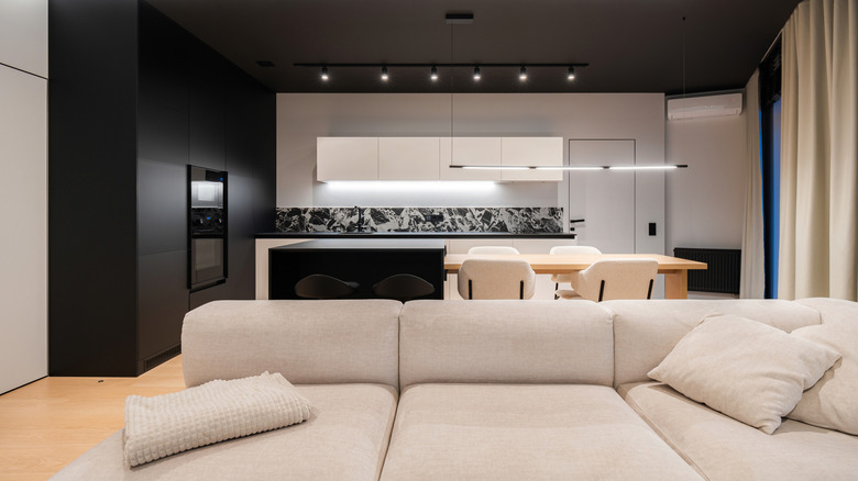 An open-concept living area with with a beige couch in the foreground and a black and white kitchen in the back