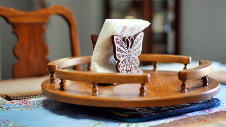 A wooden lazy Susan on a dining table contains a single napkin holder