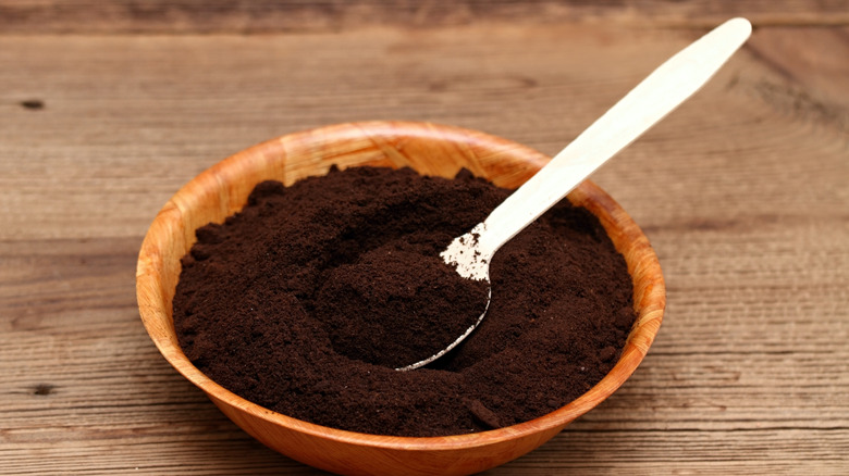 A bowl of used coffee grounds with a white plastic spoon in it sits on a wooden tabletop.