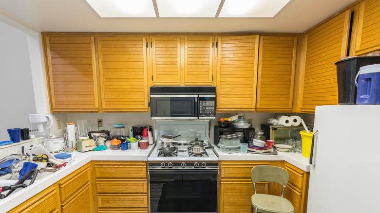 A messy, outdated kitchen with shutter-style cabinet and white tile counter tops.