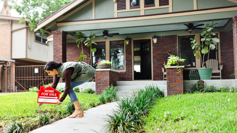 A person placing for sale sign in front of a brick home with potted plants and seating on the patio