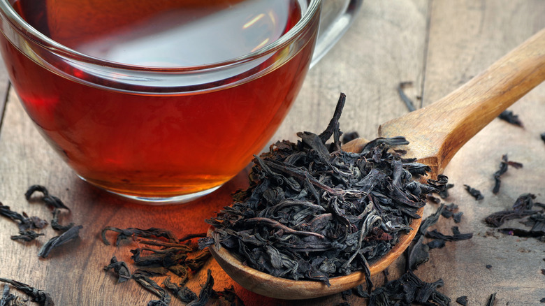 Black tea leaves in a wooden spoon near a cup of brewed tea on a wood surface