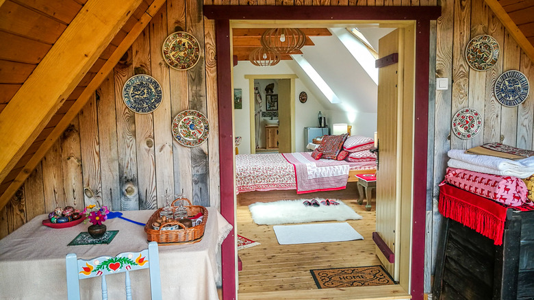 A folk art interior decor with a view from the kitchen and dining area, and a doorway that leads to a bedroom. Folk art, plates displayed on the walls, and floral bedsheets are included.
