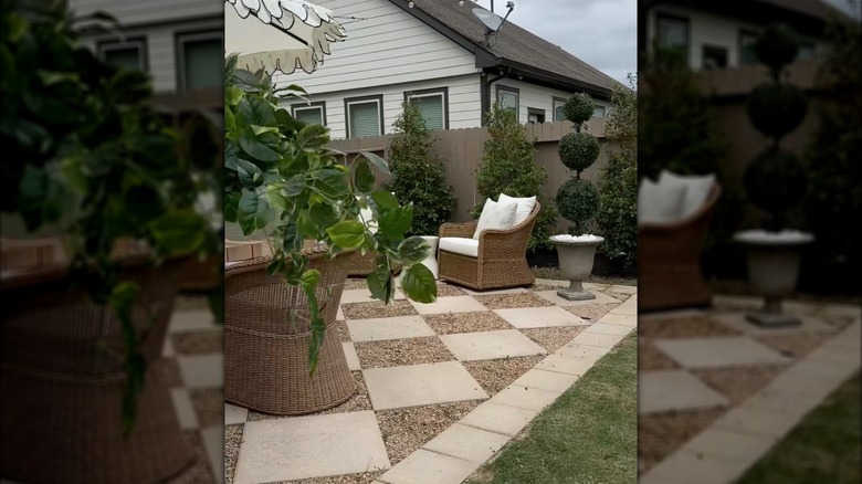 A residential backyard with a checkerboard patio, rattan outdoor furniture, and potted topiaries.