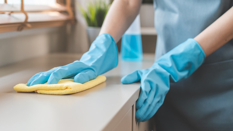 Hands in blue rubber gloves use a light yellow microfiber towel to wipe down a counter
