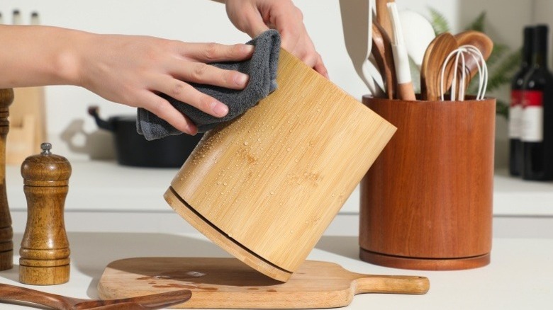 A hand wiping the outside of one of the utensil holders with a cloth while another holder sits on the bench filled with utensils