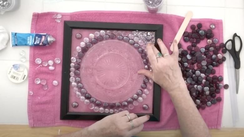 A woman attaches glass chips around a vintage plate and flat floral gems to a DIY picture frame suncatcher