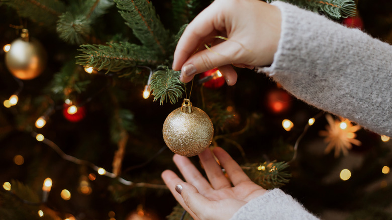 Hands hang a gold ball ornament on a lit Christmas tree.