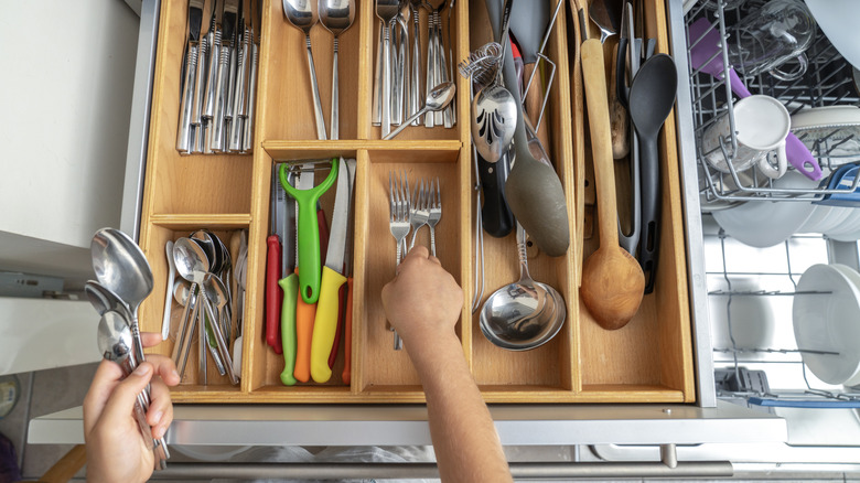 A pair of hands is putting silverware back into a wooden utensil tray that is full of silverware and serving utensils.