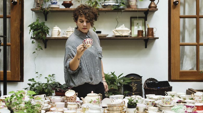 Woman looking at an old tea cup over a table full of vintage tea cups, saucers, and tea pots.