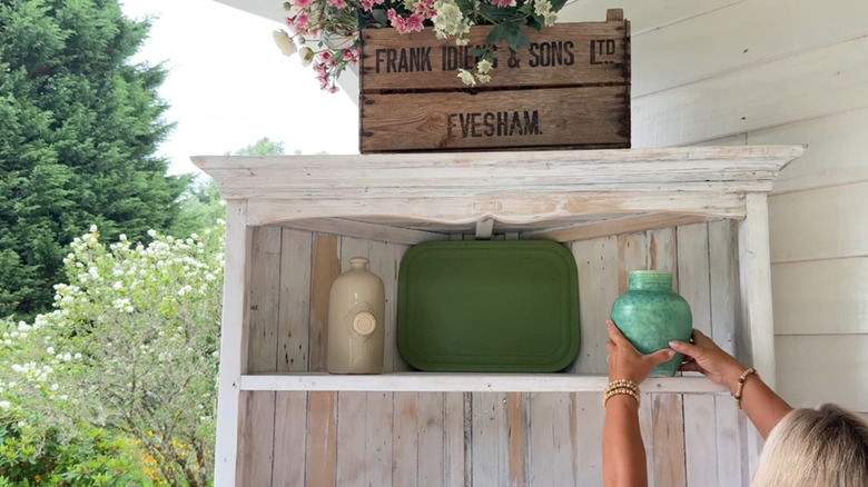 A woman adding a ceramic vase to a wooden shelf on a covered patio