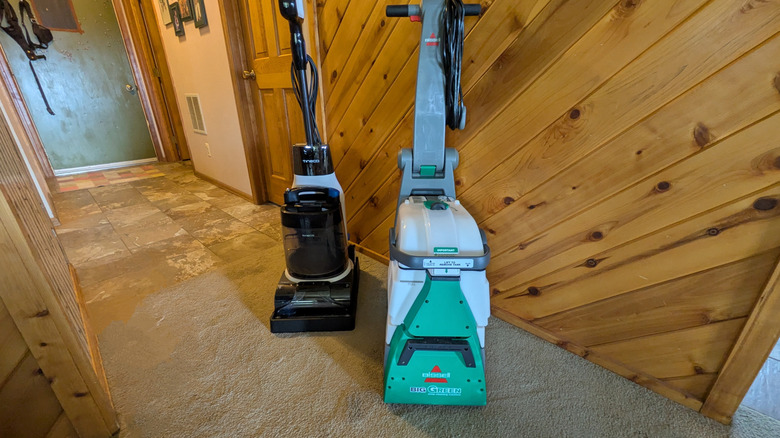 The Tineco Carpet One Cruiser and Bissell Big Green stand next to each other against a wood paneled wall.