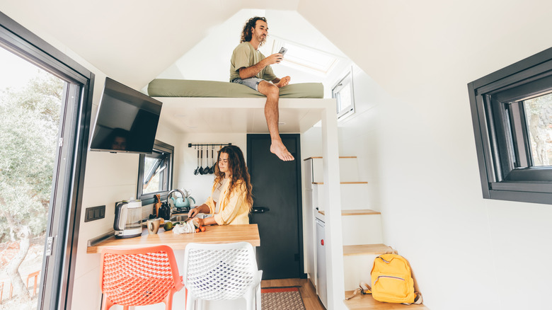 A young woman works in a kitchen while a man sits above her in the loft of a tiny home