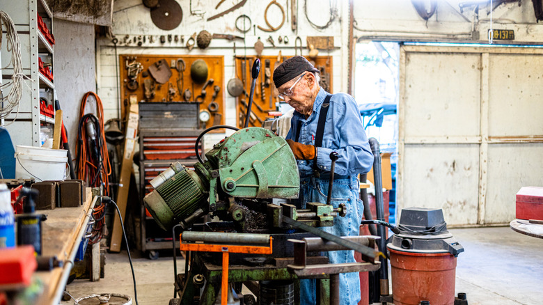 An older adult in blue overalls fixes an engine in his dedicated workshop full of vintage tools.