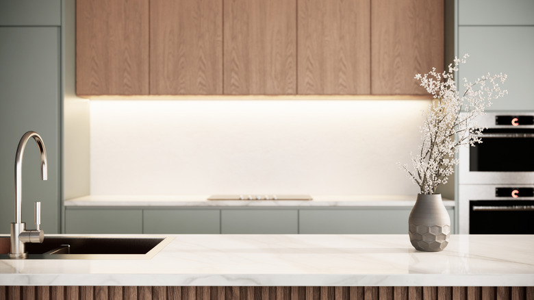 A contemporary kitchen with sage-green and natural-wood colored cabinets, out of focus with marble-topped island in foreground