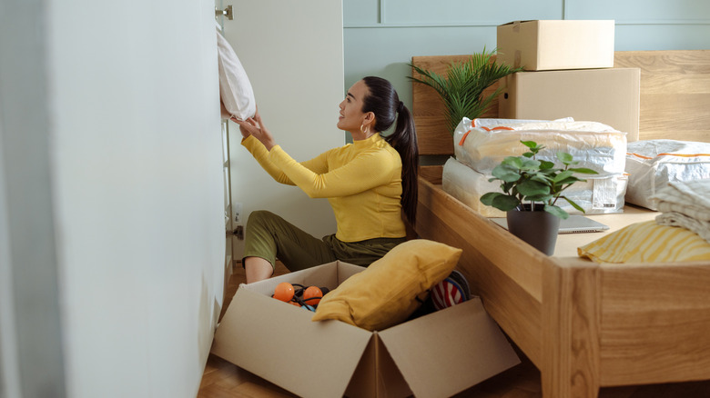 A woman sitting on a floor next to a bed and pulling items out of a closet to declutter