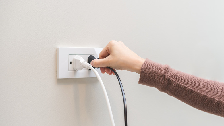 A woman in a brown shirt unplugging a cord from a white wall outlet.