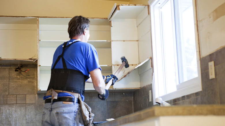 A man using a sledge hammer to tear down a kitchen cabinet insallation