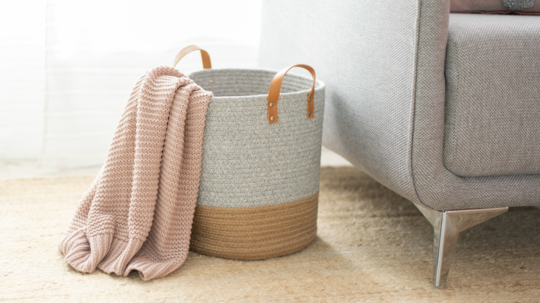 Pink knitted throw blanket draped over a woven basket with leather straps, both sit next to a grey armchair