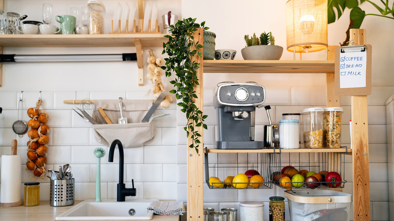 A modern kitchen with a wooden shelf that includes a coffee maker, food in jars, and two hanging fruit baskets