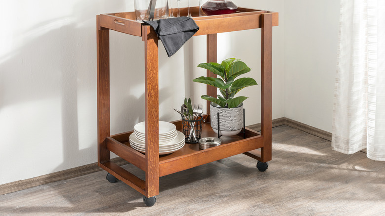 A bar cart with plates, cutlery and a house plant on the bottom shelf.