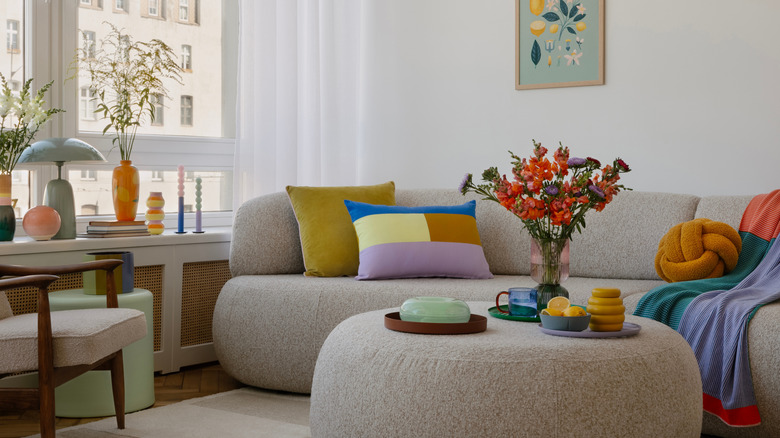 A modern curved grey sofa with matching circular ottoman in front of it with flowers in a vase and ornaments on it.