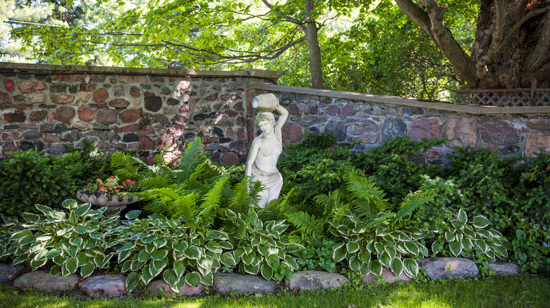 Ferns and hosta grow in a shade garden in front of a rock wall with a statue of a woman in the center