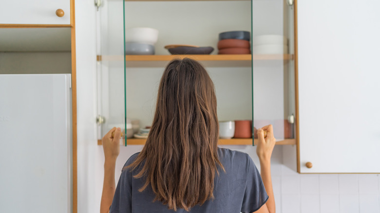 Woman opening a kitchen cabinet with plates, bowls, and mugs inside