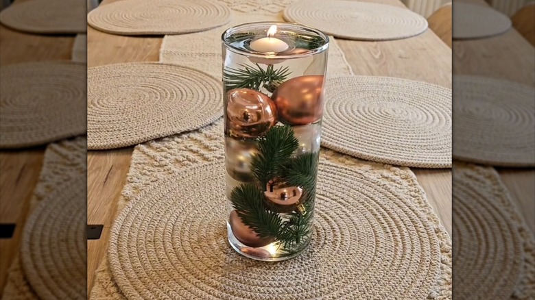 A glass vase filled with ornaments and sprigs of pine in water on a table