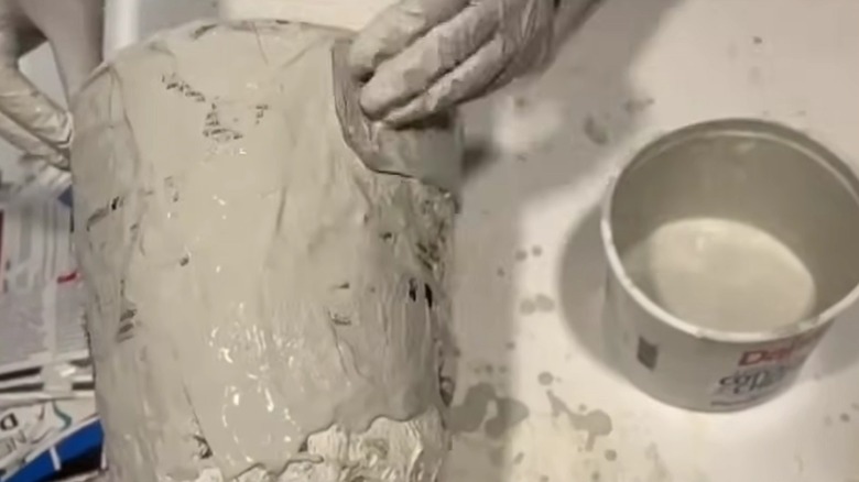A pair of gloved hands cover a plastic pot with mortar-soaked paper.