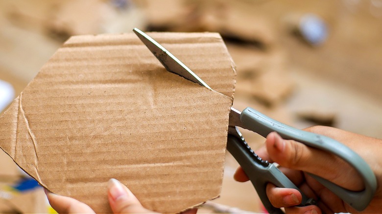 A hand holding gray scissors is cutting into a small piece of cardboard