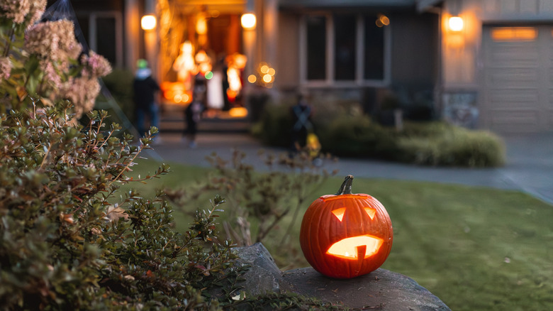 A jack-o'-lantern glows in a yard with a well-decorated porch in the background.