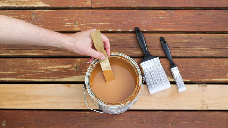 Person's hand holding a paint stir stick inside a bucket of brown paint with two paintbrushes next to it