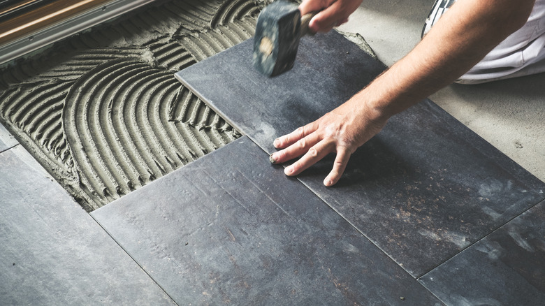 A person hammering in large dark-colored tiles in a room