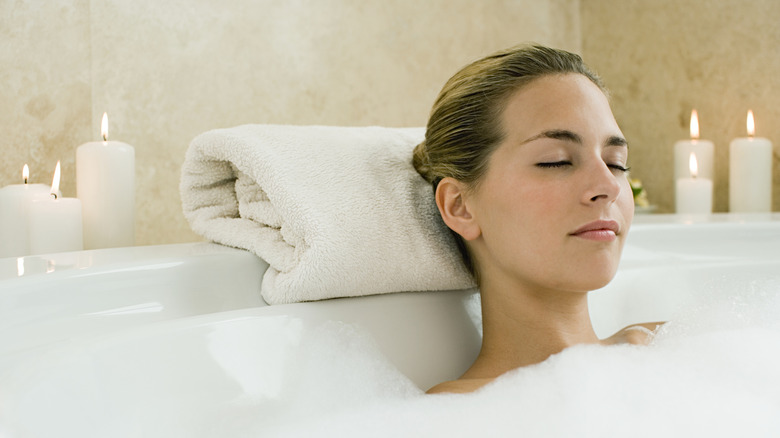 Woman relaxing in a bubble back with her head resting on a rolled-up towel and surrounded by votive candles.