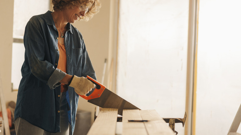 A smiling woman in a blue denim shirt using a hand saw to cut lumber.