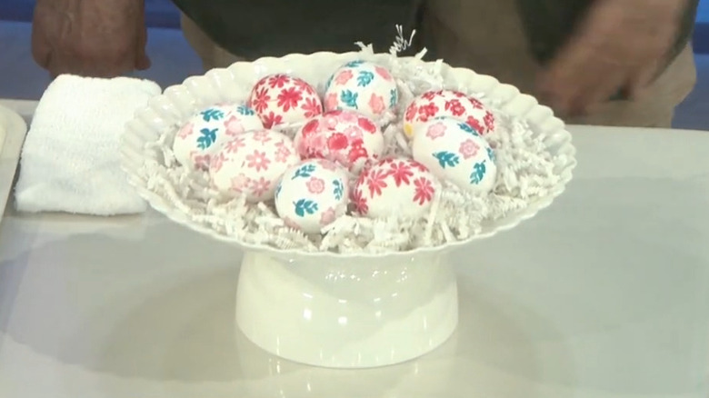 White raised bowl holds basket filling and stamped Easter eggs with floral accents