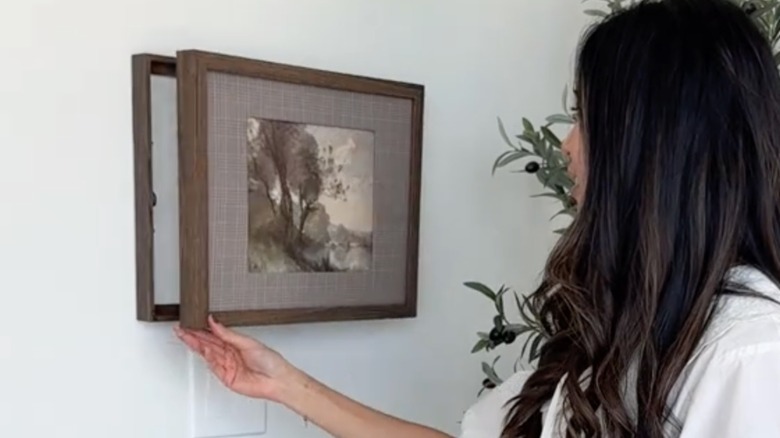 person opening hidden compartment formed from two frames attached together on wall