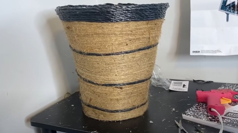 A trash can wrapped in jute and black decorative rope.