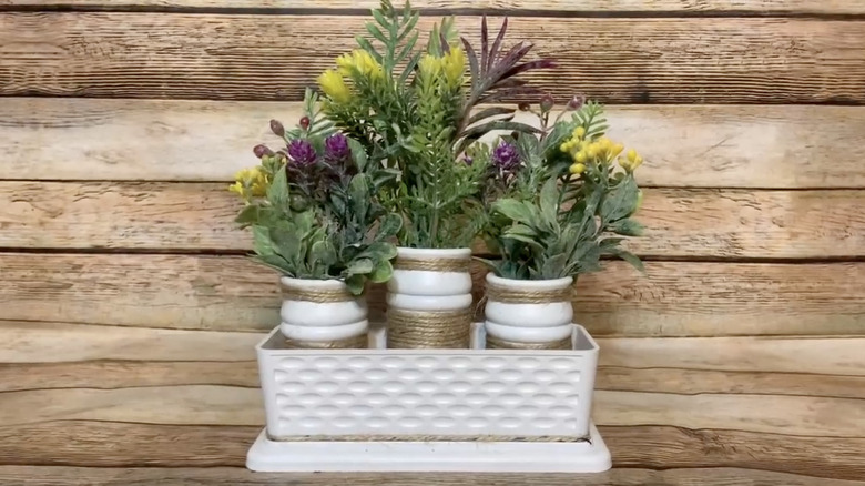 A DIY planter made from a white-painted butter dish contains three jars filled with faux plants in front of a wood backdrop
