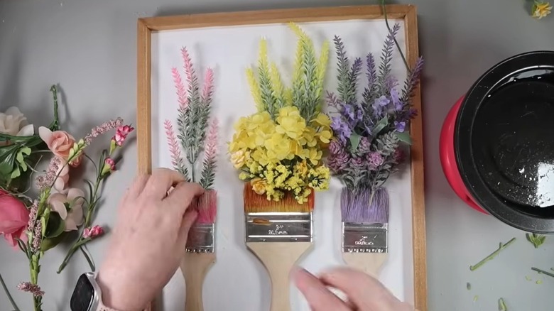 Hands gluing artificial flowers to a white canvas with paintbrushes