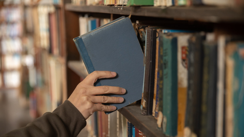 A hand takes an worn blue book from a shelf with lots of other old books.