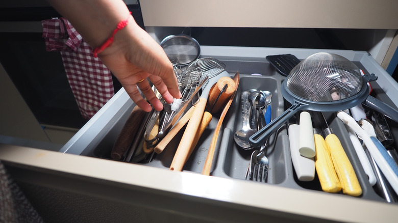 A hand reaches into a cluttered kitchen drawer full of cooking utensils and cutlery.